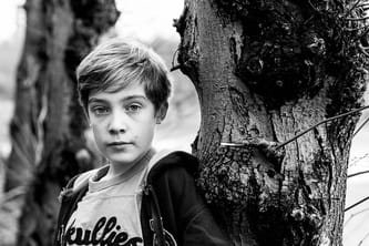 Black and white photograph of boy standing next to tree. 